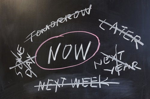 The word "NOW" with a circle around it written in the middle of a chalkboard with the words "some day", "tomorrow", "later", "next year", and "next week" all crossed out, written around the word "NOW"