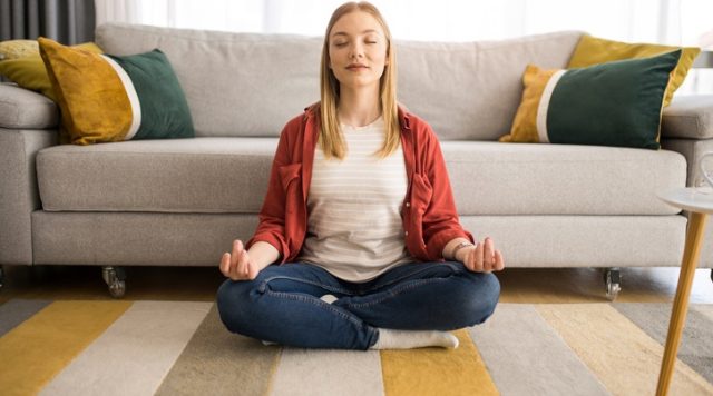 A woman sitting on the floor with her legs crossed, eyes closed, and hands resting on her knees meditating