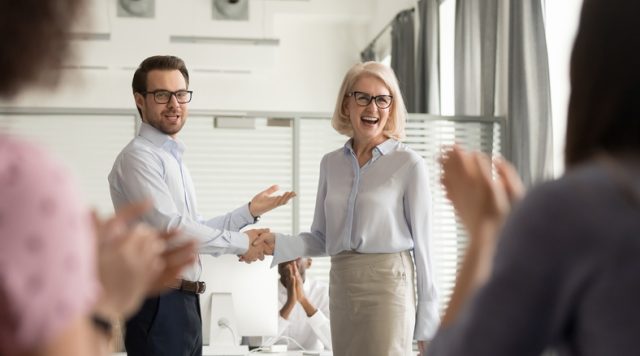 A man and a woman standing at the front of a room shaking hands. The man is pointing to the woman in acknowledgement and the rest of the people in the room are applauding
