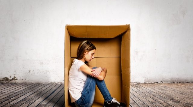 A woman sitting inside a cardboard box in the middle of the room. Her head is down and she looks sad and alone.