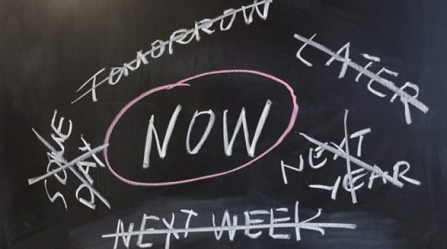 The word "NOW" with a circle around it written in the middle of a chalkboard with the words "some day", "tomorrow", "later", "next year", and "next week" all crossed out, written around the word "NOW"