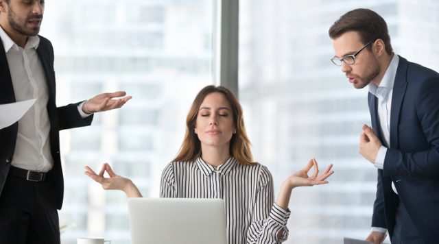Photo of a woman sitting at a desk meditating with men standing on either side of her trying to bother her