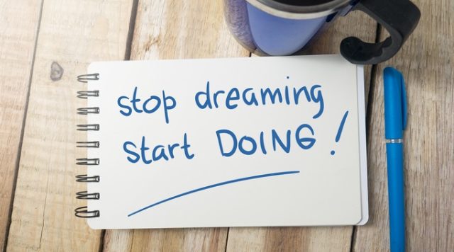 A cup of coffee, a notebook, and a pen on a table. "Stop dreaming. Start doing!" is written in the notebook